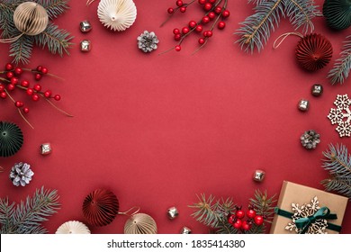 Christmas Frame With Gift Box, Paper Decorations, Spruce Branches And Berries On Red Background. Holiday Border In Earth Colours.