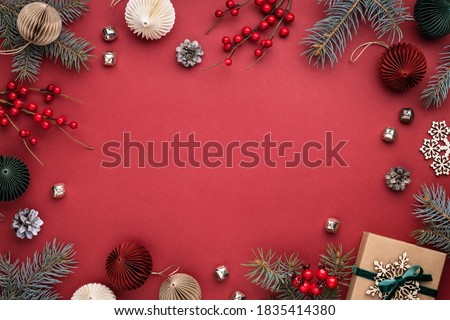 Christmas frame with gift box, paper decorations, spruce branches and berries on red background. Holiday border in earth colours.