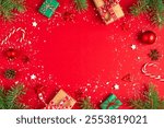 Christmas frame made of fir branches, gift boxes, festive decorations, candy and pine cones on red background. Flat lay. Happy New Year and winter holidays layout. Top view with copy space