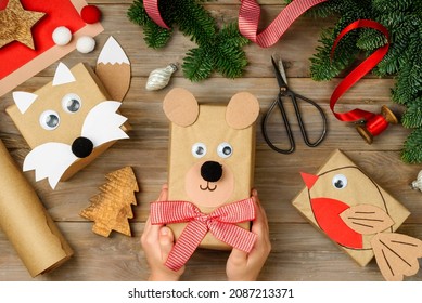 Christmas funny gifts boxes in craft paper bear, bird and fox on a wooden background.  New year   holiday concept. Festive gift wrapping. Сhildren's hands in the frame. Top view.