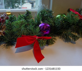 Christmas Garland With A Red Bow And Purple And Green Bobbles Sits On A Ledge Under A Window