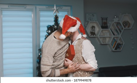 Christmas In Gay Family, Hugging In Front Of The Christmas Tree With Gifts And Santa Claus Hats. High Quality Photo