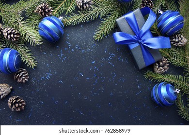 Christmas gift with blue ribbon and blue balls, tree branches and cones on dark blue background with copy space.