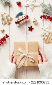 Christmas Gift In Female Hands Over White Wooden Desk With Christmas Decorations And Fir Tree Branches. Vintage Style. Flat Lay, Top View.