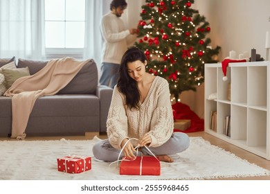 Christmas gift wrapping. An attractive mixed race Asian Caucasian woman wraps presents during Christmas and New Year preparations and a man decorates the Christmas tree. - Powered by Shutterstock