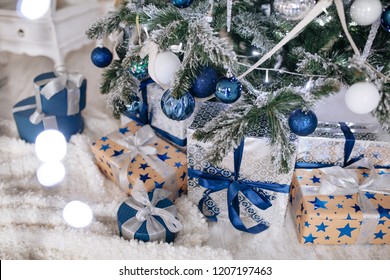 Christmas Gifts Wrapped In Silver And Blue Paper, Background With Xmas Lights Bokeh Of Blurred Under Christmas Tree.