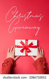 Christmas giveaway concept. Woman hand in sweater holds a Christmas gift on red background. Christmas giveaway gift - Shutterstock ID 2086710052