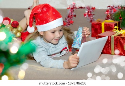 Christmas greetings online. Smiling blonde girl uses her mobile tablet to communicate. Shows gifts (online purchases) to the camera. Greeting video calls to friends, relatives, and parents