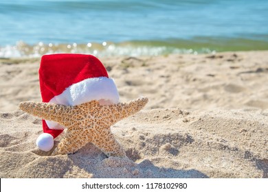 Christmas Hat On Starfish In Beach Sand With Ocean Water Background