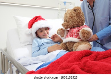 Christmas Holiday In Hospital Happy Child Lying In Bed With Santa Claus Hat And Nurse Dressing A Teddy Bear 