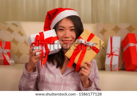 Christmas holiday lifestyle portrait at home of young beautiful and happy Asian Korean woman in pajamas and Santa hat  holding lot of present and gift boxes smiling on living room couch excited 