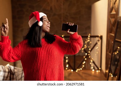 Christmas Holiday Music. Happy Black Lady In Santa Hat Singing, Using Smartphone As Mic At Home, Mockup For Mobile App. Cheerful Young Woman Enjoying Karaoke Song, Having Fun On Xmas