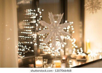 Christmas And Holidays Concept - Paper Snowflake Decoration Hanging Over Garland Lights And Candles At Night Window