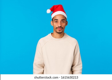 Christmas, Holidays And Family Concept. Cute And Silly African-american Guy Celebrate Winter Holidays Parents House, Wearing Santa Hat Feeling Awkward, Attend New Year Party, Blue Background