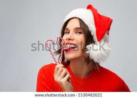 christmas, holidays and people concept - happy smiling young woman in santa helper hat licking candy canes over grey background