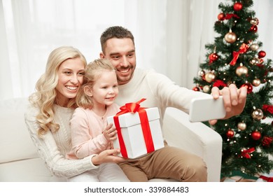 Christmas, Holidays, Technology And People Concept - Happy Family Sitting On Sofa And Taking Selfie Picture With Smartphone At Home