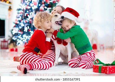 Christmas At Home. Kids And Dog Under Xmas Tree. Little Boy And Girl Hug Pet In Santa Hat And Open Christmas Presents. Children Play With Animal. Winter Holiday Celebration. Love And Friendship.