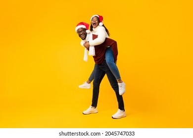 Christmas Joy. African American Family Couple Posing Wearing Santa Hats Having Fun, Husband Carrying Wife Piggyback Smiling To Camera On Yellow Studio Background. Merry Xmas Concept. Full Length - Powered by Shutterstock