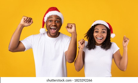 Christmas Joy. Black Couple In Santa Hats Shaking Fists In Excitement, Celebrating Winter Holidays Standing Over Yellow Studio Background. New Year And Xmas Celebration Concept. Panorama