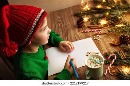 Christmas Kid writing Letter to Santa. Happy Child in Red Santa Hat dreaming about Presents at Home. Xmas Gifts Shopping Wish List over brown Wooden Table decorated with Christmas Lights, Fir Tree - Powered by Shutterstock