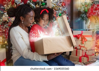 Christmas Magic. African American Mom And Daughter Opening Shining Christmas Gift Box, Sitting Near Xmas Tree, Celebrating Holidays At Home