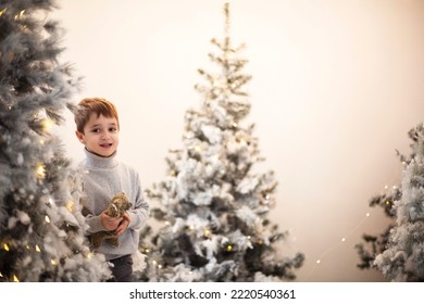 Christmas Miracle. Cute Happy Little Boy With Teddy Bear Surrounded With Snow-covered Artificial Xmas Trees Decorated With Yellow Twinkle Lights, Selective Focus. Children At Christmastime