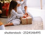 Christmas morning in pajamas. A mother with a baby and an older child are holding gifts in their hands. A Christmas tree in the background.