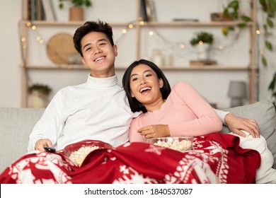 Christmas Movies Night. Happy Chinese Couple Watching Movie On TV Holding Television Remote Control, Eating Popcorn Sitting On Couch At Home, Covered With Cozy Winter Blanket. Family Enjoying Xmas Eve