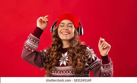 Christmas Music. Pretty Young Woman In Headphones And Winter Outfit Dancing To Her Favorite Song On Red Studio Background, Panorama. Lovely Lady Enjoying Xmas Holiday Playlist