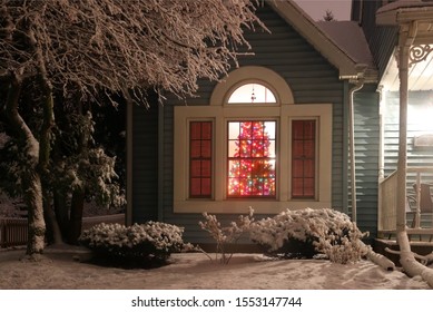 Christmas And New Year Background. Night Scene With Decorated Christmas Tree Glowing In A Room Of The Private House, View From Outdoor With Fresh Snow On A Front Yard In Foreground.