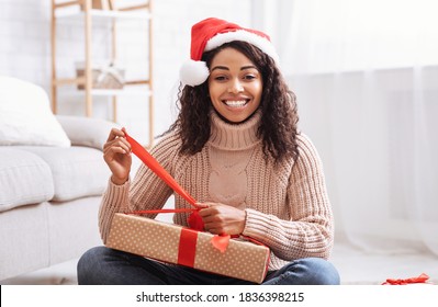 Christmas And New Year Celebration. Excited Black Woman Opening Xmas Present And Looking At Camera. Smiling Lady Unwrapping Gift Box With Red Bow, Sitting On The Floor At Home In Living Room At Home