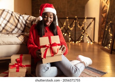 Christmas and New Year celebration. Excited young black woman in Santa hat and warm sweater opening Xmas gifts on floor at home, copy space. Millennial lady unboxing holiday presents indoors - Powered by Shutterstock