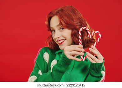 Christmas and New Year! A cute cheerful girl with bright red hair in bright green clothes holds christmas candy canes and smiles. Christmas joy. Red background. - Powered by Shutterstock