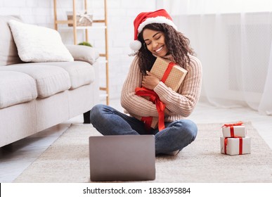 Christmas And New Year Presents. Happy Black Lady Holding And Embracing Gift Box, Sitting On The Floor At Home In Living Room With Laptop Computer, Celebrating Online, Making Video Conference - Powered by Shutterstock