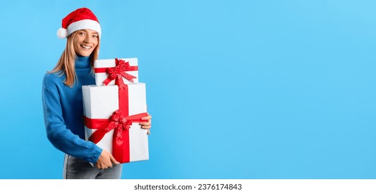 Christmas, New Year sale. Happy lady in Santa hat holding gift boxes and smiling at camera on blue studio background, panorama, free space. X-mas celebration, winter holidays mood - Powered by Shutterstock