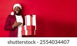 Christmas, New Year and shopping concept. Cheerful Black man secret santa holding xmas presents and smiling excited, bring gifts, standing against red background.