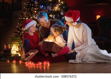 Christmas Night. Near The Christmas Tree A Lovely Family Opening Their Gifts. They Enjoy The Warm Christmas Atmosphere In Their Living Room, Mom And Kids Wearing A Hat Of Santa Claus.