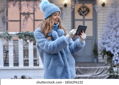 Christmas online greetings during coronavirus.A beautiful girl greets, congratulates, talks using a tablet for video calls to friends,family or takes a selfie on the background of the Christmas house. - Shutterstock ID 1856296132