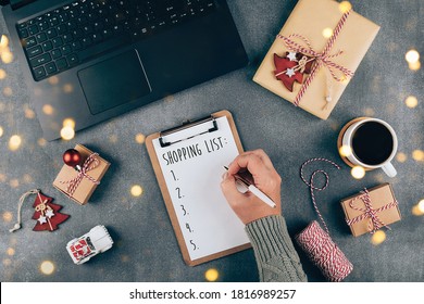 Christmas online shopping flat lay. Girl writing shopping list. Laptop, present box, cup of coffee, holiday decoration. Winter holidays sales