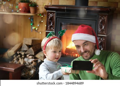 Christmas Online Video Chatting, Virtual Greetings, Giving A Gift. Father And Son Congratulations Family And Friends Using Smartphone. New Year Home Celebration.