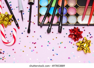 Christmas Party Makeup. Bright Color Glitter Eyeshadow, Mascara, Eyeliner, Red Lipstick, Lip Liners, Brushes And Applicator With Candy Cane, Gift Wrap Bows And Confetti On Pink Background. Copy Space