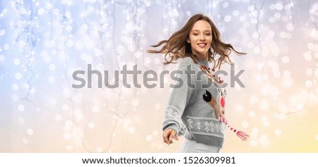 christmas, people and holidays concept - happy young woman in jumper with reindeer pattern dancing at ugly sweater party over festive lights background