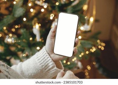 Christmas phone mock up. Hand holding smartphone with empty screen against stylish festive christmas tree with golden lights. Space for text. Christmas advertising, smartphone app template - Powered by Shutterstock