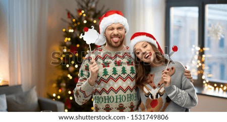 christmas, photo booth and holidays concept - happy couple in ugly sweaters posing with party props over home background
