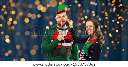 christmas, photo booth and holidays concept - happy couple in ugly sweaters posing with party props over festive lights on dark night background