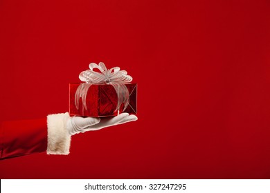 Christmas. Photo Of Santa Claus Gloved Hand With Red Gift Box, On A Red Background