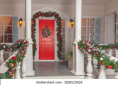 Christmas porch decoration idea. House entrance with red door decorated for holidays. Red and green wreath garland of fir tree branches and lights on railing. Christmas eve at home. - Powered by Shutterstock