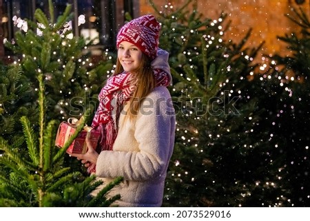 Christmas portrait of a beautiful girl in a warm scarf, standing on the street, decorated for the New Year, against the background of a Christmas tree with a gift in her hands
