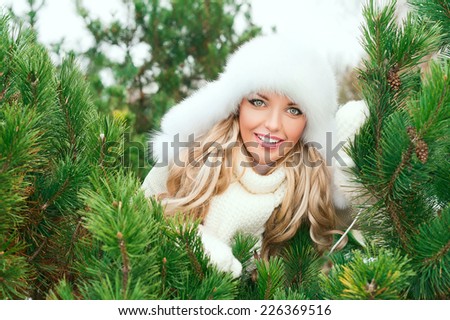 Christmas poster. Young smiling woman with fur white hat, woolen mittens, sweaters in the winter fir forest. Fir branches.