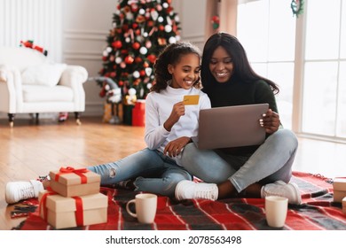 Christmas Preparation. Smiling black girl and lady using pc notebook, choosing, ordering and buying gifts online, cheerful teen daughter holding golden credit card in decorated living room interior - Powered by Shutterstock